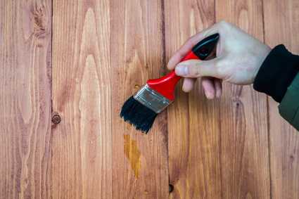 Holzlasur: Eine Hand hält einen roten Pinsel und trägt damit Farben-Beize auf eine Holzoberfläche auf. - Boden Pelzer -Bodenbeläge, Vinylbelag, Parkett - Moselstr. 14 - 41464 Neuss