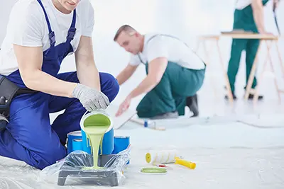 Zwei Maler bereiten sich auf Malerarbeiten vor; einer gießt grüne Farbe in eine Schale, während ein anderer im Hintergrund arbeitet. Farbvorräte sind auf einem mit Plastik bedeckten Boden verstreut. - Firmenadresse