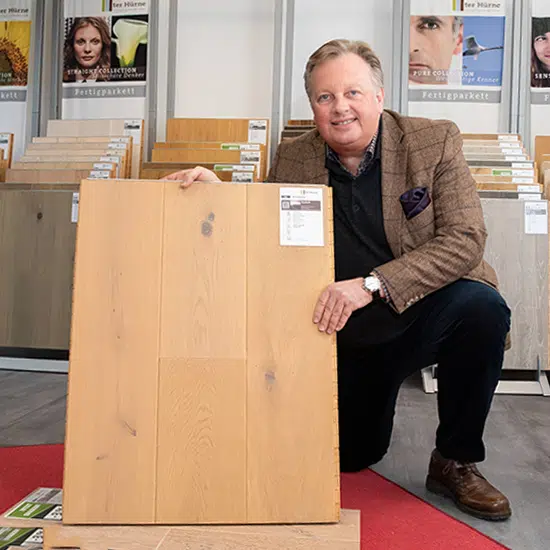 Hans Peter Pelzer kniet neben einem großen Holzbodenmuster im Ausstellungsraum von Boden Pelzer, im Hintergrund sind mehrere Bodenbelagsdisplays elegant angeordnet. - Boden Pelzer -Bodenbeläge, Vinylbelag, Parkett - Moselstr. 14 - 41464 Neuss