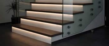 Holztreppe mit Glasgeländer, hervorgehoben durch LED-Beleuchtung unter der Stufe. Links auf dem dunklen Fliesenboden steht eine Topfpflanze. - Fussbodentechnik Schwallach -Bodenbeläge, Vinylbelag, Parkett - Neckaraue 7 - 71686 Remseck